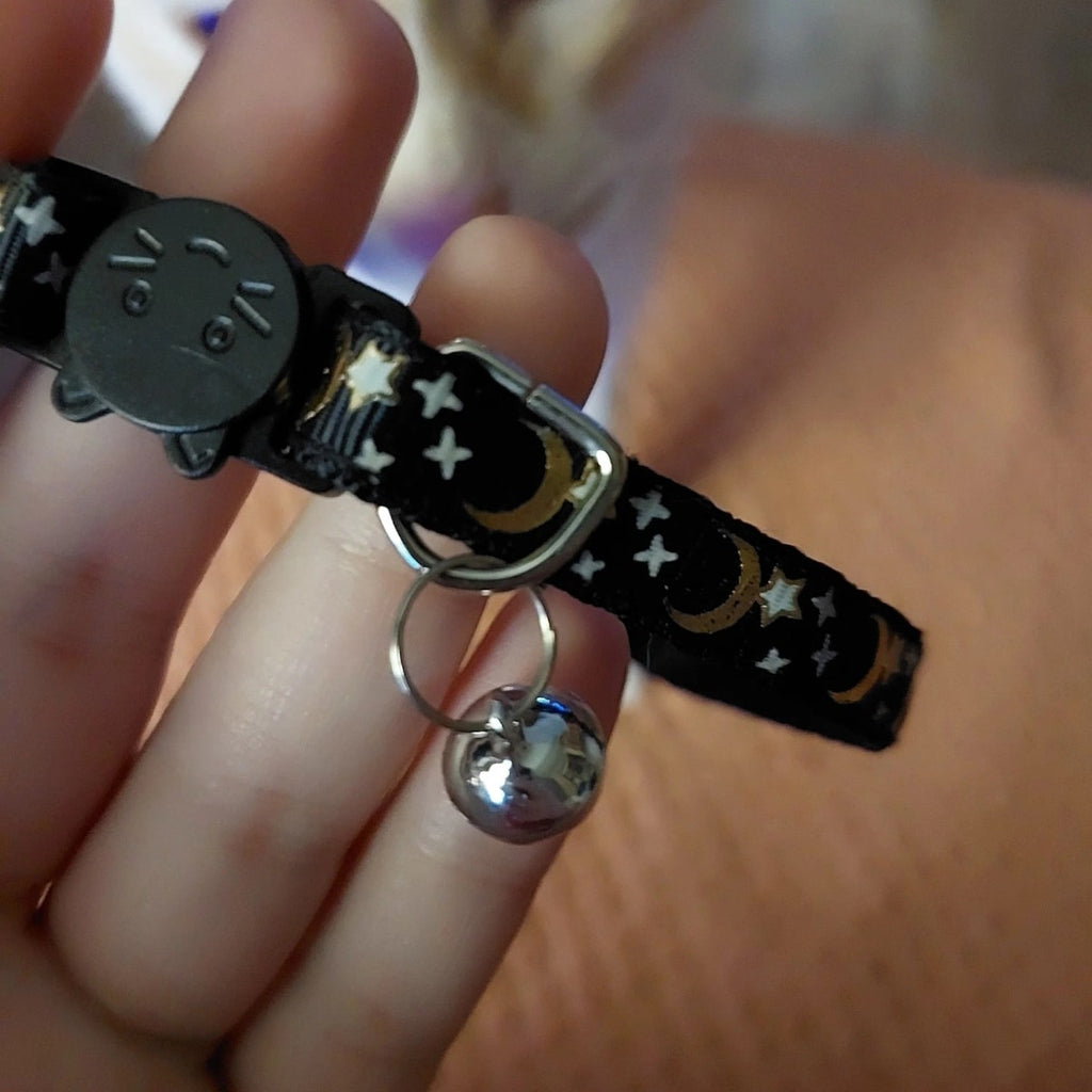Black cat collar held on two fingers, showing the cat-face buckle, the D-ring and the silver bell on its split ring