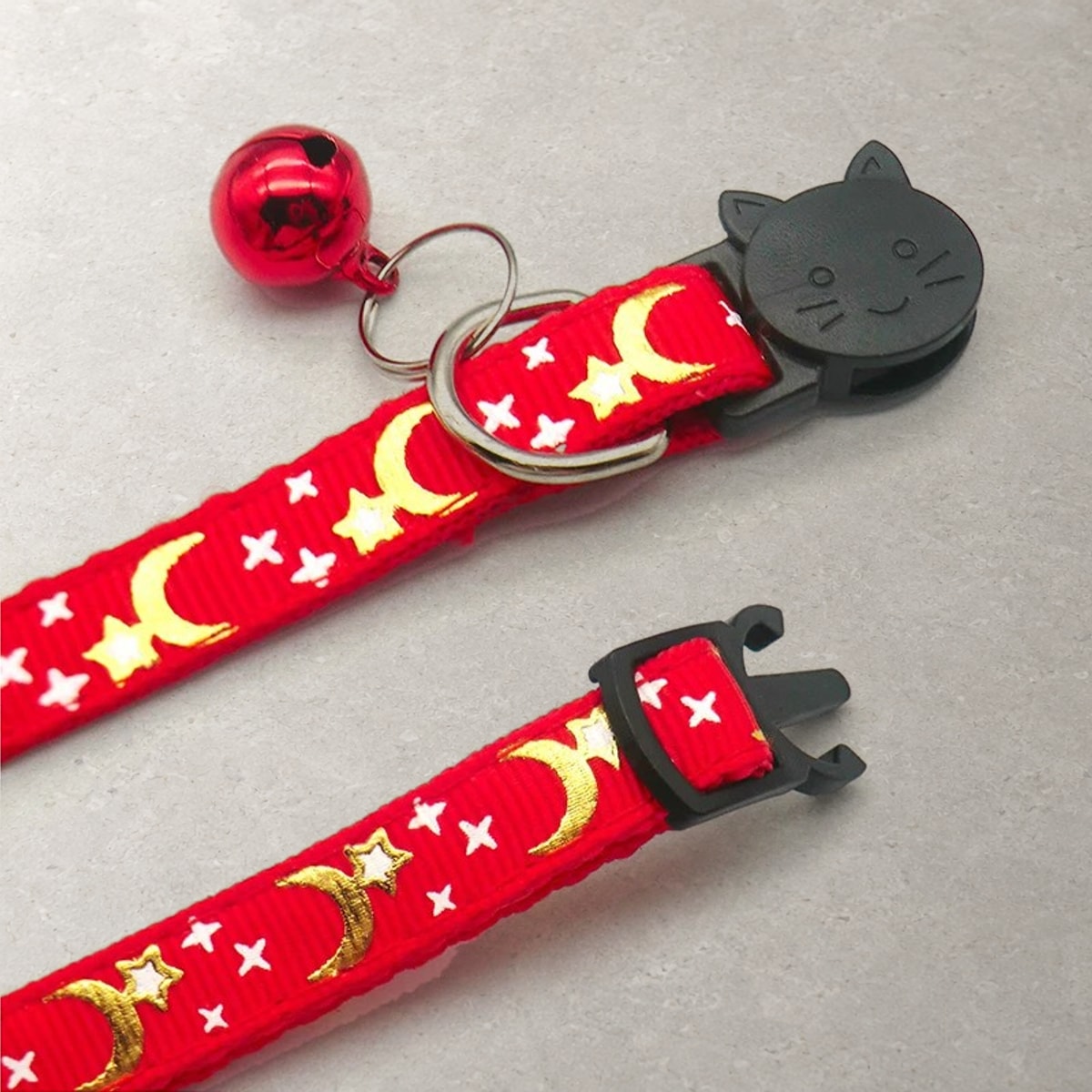 Red breakaway cat collar with its cat-face safety buckle pulled apart, beside the D-ring and the bell on its split ring